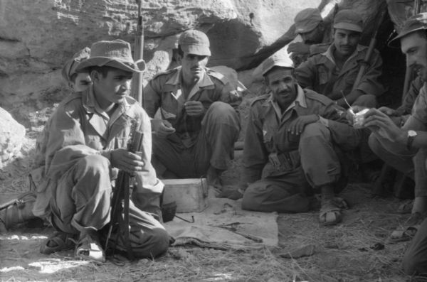 A group of Algerian National Liberation Front members sitting watching another member holding a can and what appears to be a fuse. The men are dressed in fatigues and are holding rifles.