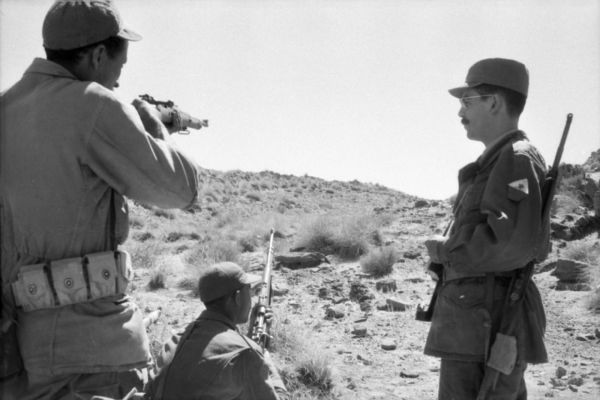 Algerian National Liberation Front members firing off their weapons into the desert. One man is crouching in front of another, while another man is watching them on the right.
