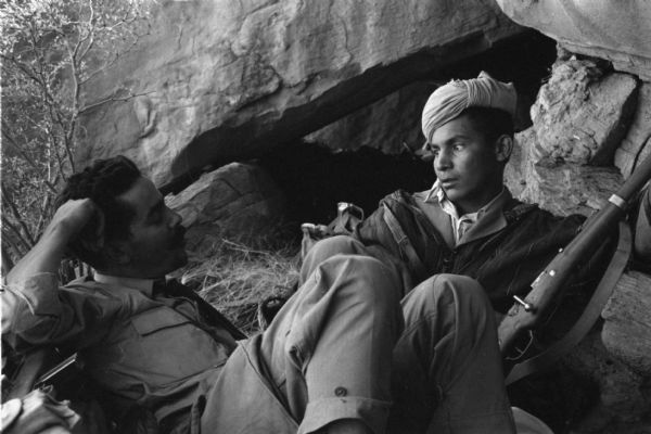 Two National Liberation Front members sit and talk outside of a cave. One man in a turban is holding a rifle.