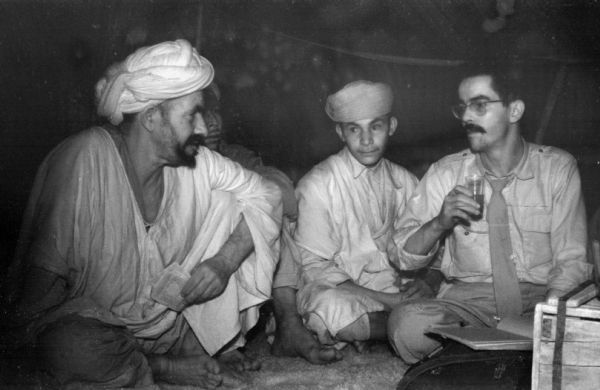 Algerian National Liberation Front members gather for a meeting while sitting on the floor of a tent. Two men are wearing turbans, one of them holding paper bills. The man on the right is wearing a tie and is holding a glass in his hand.
