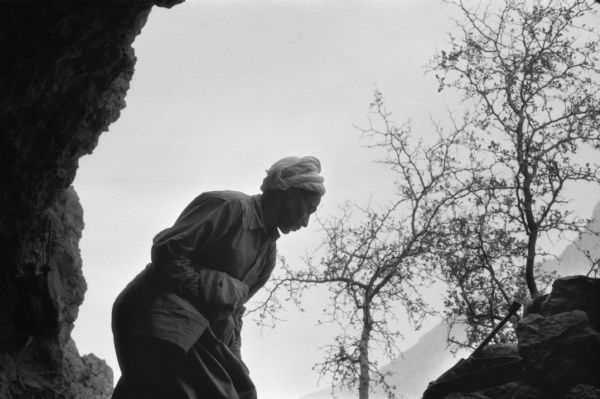 View from inside of cave of an Algerian member for the National Liberation Front in the Algerian desert. He is wearing a turban, and in front of him resting against a rock is a rifle. Behind him are trees and mountains.