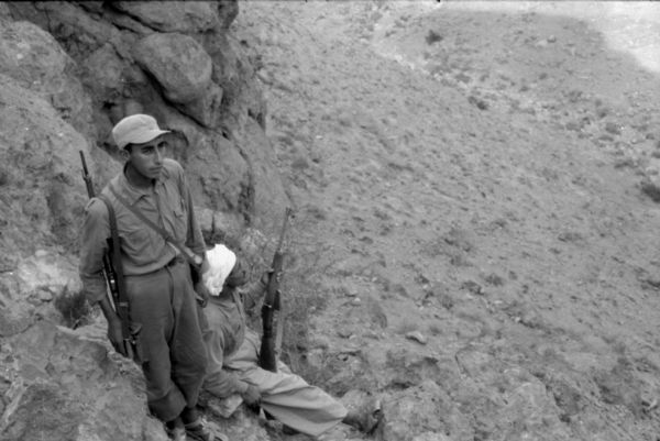 Two Algerian members of the National Liberation Front on patrol around their camp. One man is standing on top of rocks and the other man is sitting on the rocks. They both have rifles.