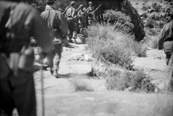 Algerian members of the National Liberation Front marching to line up in front of other FLN members in the desert. They are all wearing fatigues and carrying rifles. There are shrubs in the foreground.