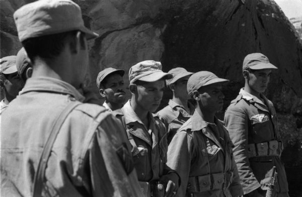 An Algerian group of National Liberation Front members gathered in a group. They are wearing caps and fatigues and some of them are standing at attention. One of them has a grenade strapped to his belt and another is holding a rifle.