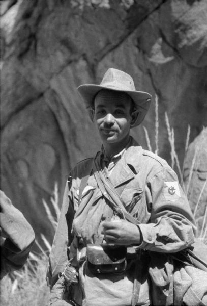 An Algerian National Liberation Front member posing for a photograph outdoors. He is wearing a hat, and fatigues with the FLN insignia on the sleeve. He has several bags slung over his arm and a grenade attached to his belt.