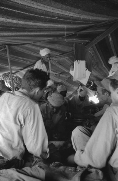 A group of Algerian National Liberation Front members gather in a tent for a meeting. Some of the men are wearing turbans and some are wearing caps. They are sitting in a circle eating. The fabric of the tent is striped.