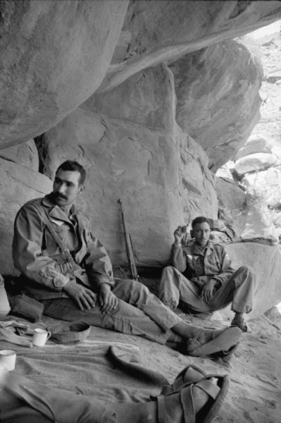 Algerian members of the National Liberation Front sitting at the entrance of a cave having tea. Rifles are leaning against the rocks behind them. The men are wearing fatigues and reclining against the boulders.