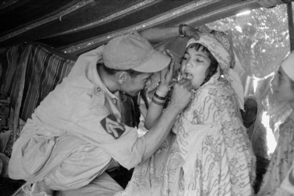 An Algerian Red Crescent (Red Cross equivalent) examining a young girl's teeth. The girl is wearing a patterned shawl and is reaching for the man's hat. They are inside a Red Crescent tent, and two other people are sitting on the right.