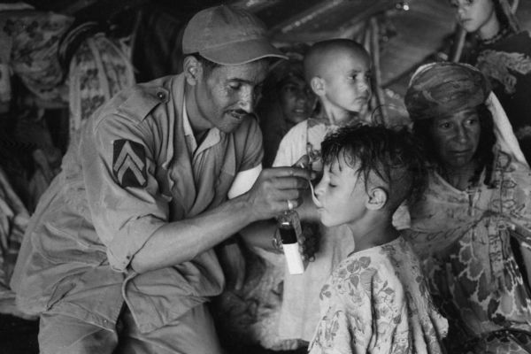 A Red Crescent (Red Cross equivalent) worker spooning medicine into a young child's mouth. The child's head is half shaven. Behind them are two women, a young child, and an older girl. They are inside a Red Crescent medical tent.