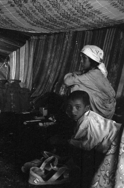 A young Algerian boy and girl waiting side-by-side in a Red Crescent (Red Cross equivalent) tent. Beside the boy are a pair of sandals. The girl is wearing a head scarf. The tent is made of striped and flowered fabric.