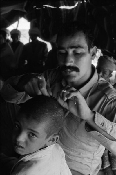 An Algerian member of the National Liberation Front cutting his young son's hair in a tent. Behind him is a group of men.