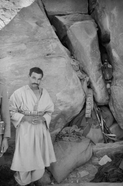 An Algerian member of the National Liberation Front posing by a cave wall. Behind him are large canvas bags, guns, and clothing stored between large boulders.