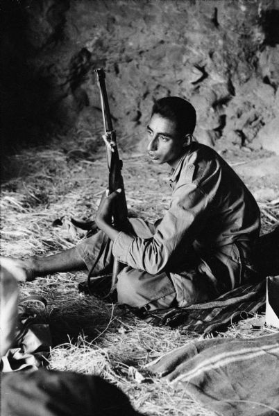 An Algerian National Liberation Front member sits in a cave with his rifle. He is sitting on a striped blanket.
