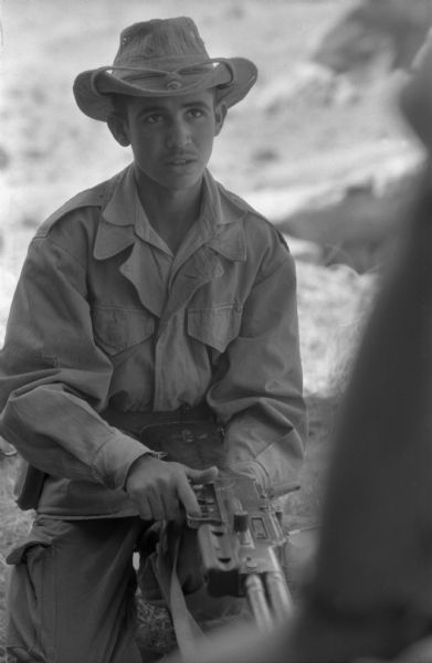 A young Algerian National Liberation Front member listening to another member speak. He is holding a large gun and is wearing a hat. Behind him is the Algerian desert.