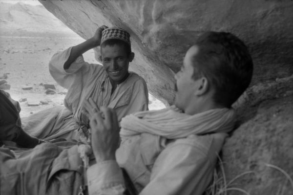 Two Algerian National Liberation Front members relax and laugh together near the entrance of a cave. In the background is the desert.
