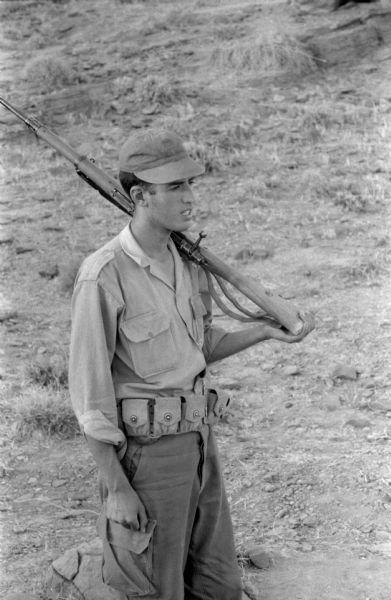 An Algerian member of the National Liberation Front standsingoutdoors in the Algerian desert with a rifle over his shoulder. He is wearing a cap and fatigues.