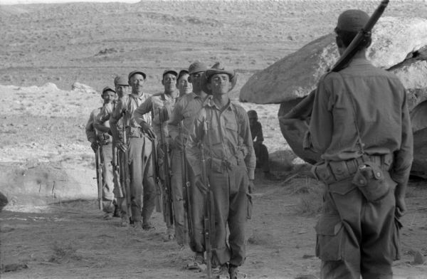 A group of National Liberation Front members stand at attention in front of another member with a rifle on his shoulder. The men are preparing for training. The Algerian desert is behind them.