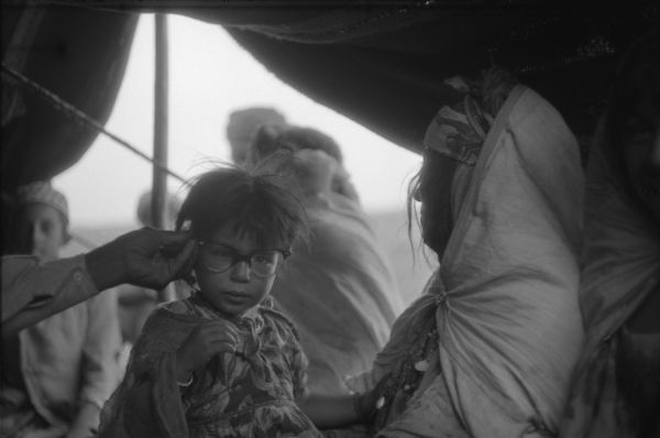 An Algerian girl in a Red Crescent (Red Cross equivalent) tent. A man is holding Dickey Chapelle's glasses up to the girl's face. She is wearing a patterned dress and is being held by a woman in a shawl. Behind them are other people and the opening of the tent.
