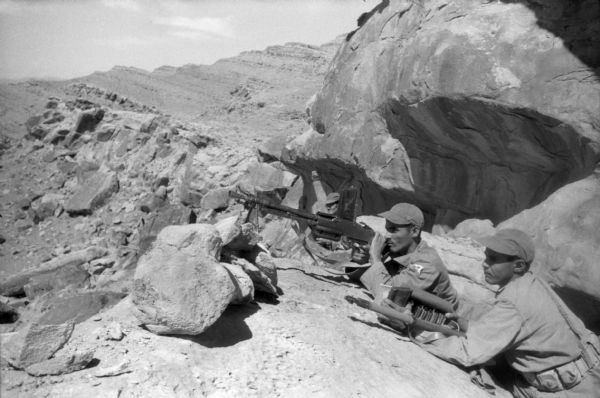 Algerian National Liberation Front members hold lookout positions with machine guns. They are wearing fatigues and behind them is the rocky Algerian desert.