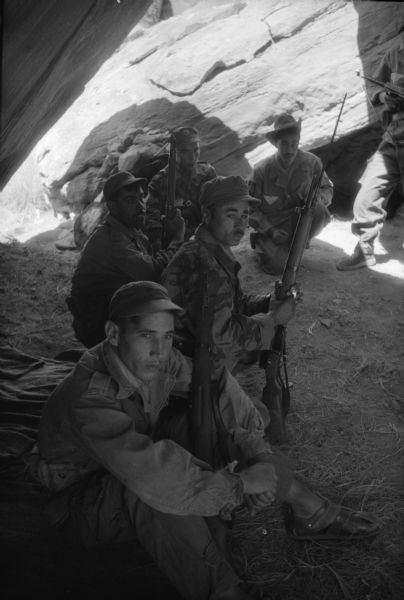 A group of Algerian National Liberation Front members sitting in a cave with their rifles. They are wearing fatigues.
