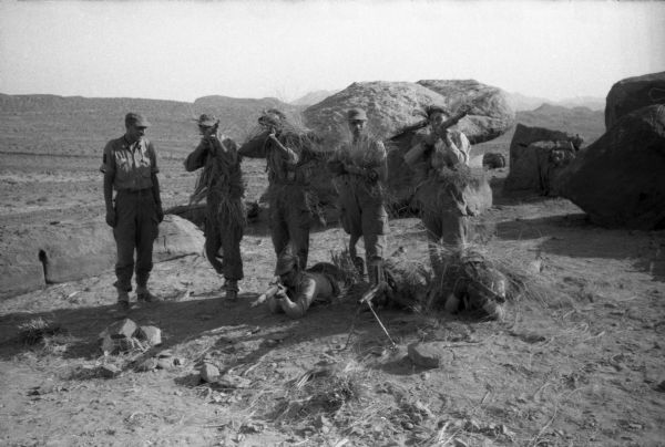 A group of National Liberation Front members posing for a photograph. They are wearing long grasses for camouflage and holding rifles and submachine guns. Behind them is the Algerian desert.