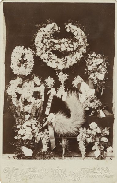 Funeral memorial card of floral wreaths with a banner reading, "AT REST," and an animal tail [?].