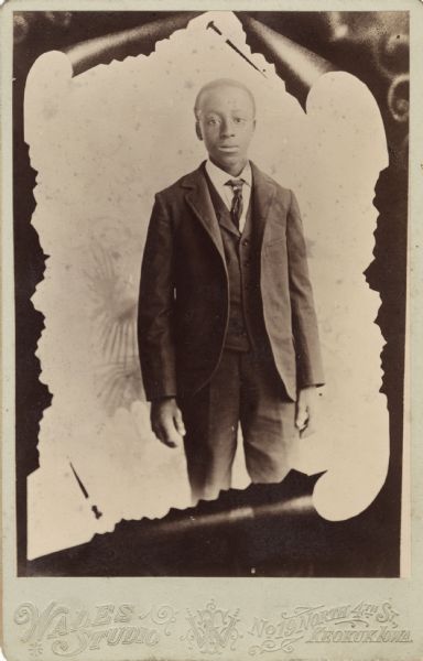 Studio portrait of a three-quarter length view of an African American youth wearing a three-piece suit and a tie, in front of a painted backdrop. The image is in the form of a pinned-up scrap of paper.