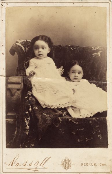 Two young girls in white dresses trimmed with lace posing for a studio portrait on a bench covered with a kilim rug.