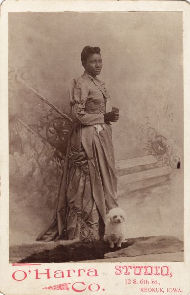 Woman in a long dress wearing a glove and holding a book [?] in her hand posing before a painted backdrop. There is a small white dog seated on a fur rug at her feet.