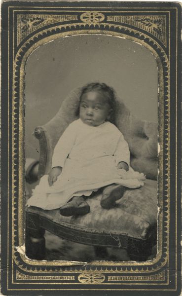 Tintype/ferrotype of a child sitting on a bench wearing a white dress and shoes.