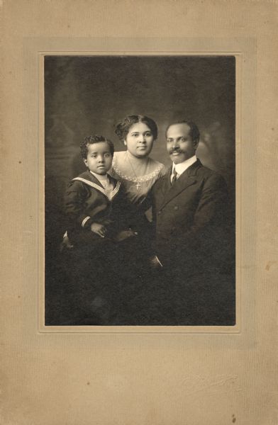 Studio portrait of an African American family (probably Caroline Webb, Andrew Norris Webb Sr., and Andrew Norris Webb Jr.) in front of a painted backdrop. The man is wearing a double-breasted suit, shirt, and tie, the woman is wearing a sheer-yoked dress and a cross pendant, and the boy is wearing a sailor suit.