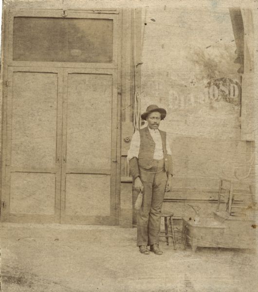 An African American man wearing a hat, shirt, open vest, trousers, and boots, posing by a shoeshine stand in front of a commercial building.