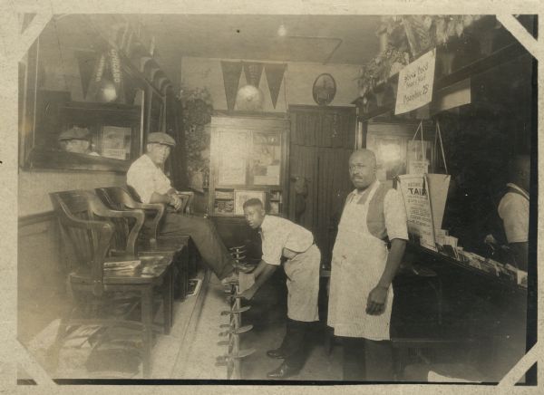 Two African American men working in a shoeshine shop. The younger man is shining the shoes of a white customer who is sitting in one of the chairs on the elevated stand, while an older man stands off to the side. All three men are looking at the camera. Banners reading "Chicago," "Iowa," and "Wis[consin]" hang from the walls of the shop.