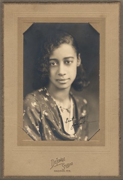 Quarter-length studio portrait of a young African American woman wearing a cowl-necked flowered dress and a necklace. The image is inscribed, "Lest you forget—".