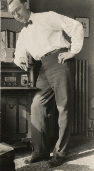 Newspaperman Alvin Steinkopf leaning on a large radio in his Milwaukee apartment. Steinkopf is wearing a bow tie and holding a cigar in his hand.