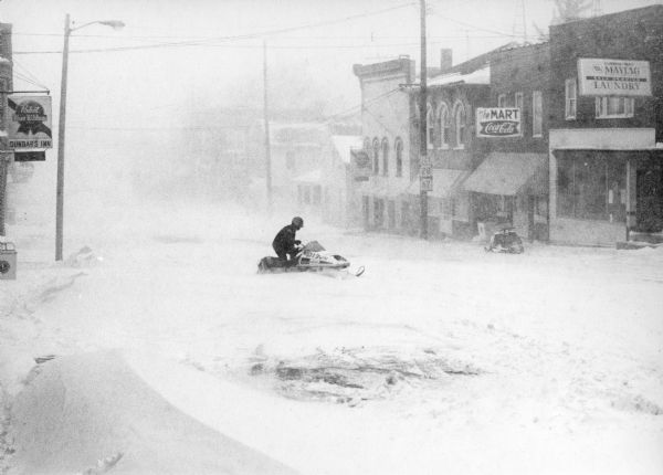 Winter scene with Kathy Thompson riding her snowmobile at the height of a snowstorm, through downtown Theresa, Wisconsin. "The village was victim of an old fashioned, swirling, snowstorm. Huge drifts made many roads impassable. Due to the fact that February proved to be a very cold month, most of the snow remained on the ground during that period."