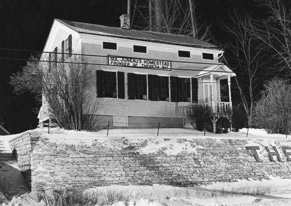 "Sol. Juneau's Homestead at night. In June, 1932, the house was moved to this location from its original site, higher up on the hill." Sign on residence reads, "Founder of Theresa, 1847."