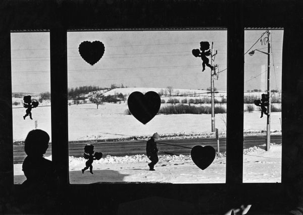"With the approach of Valentine's Day, the camera looks through the decorated windows of the Bob Bernhard home. Brenda watches her brother, Scott, playing in the snow."