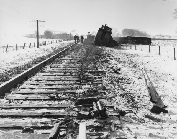View of a people walking on railroad tracks near a derailed train.