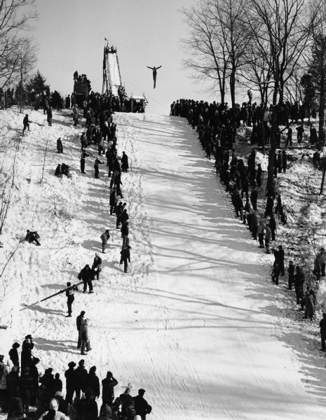 A man jumps off a ski ramp as many spectators look on.