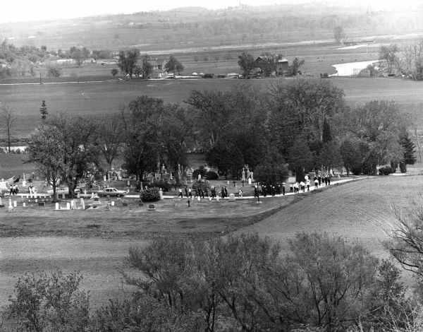 View of a parade traveling through a cemetery. Beyond, barns and farmhouses can be seen.