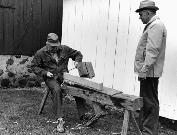 "Ist das nicht ein schnitzel bank? Ja das ist ein schnitzel bank. This bench made by Aug. Beck (1951-1910) used to hold wood so it can be whittled with a draw knife. Grandsons, Alphonse and Les Beck demonstrate."