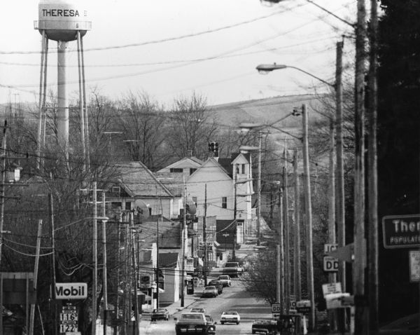 View of automobiles driving on a street, entering the city. The Theresa water tower can be seen on the left.