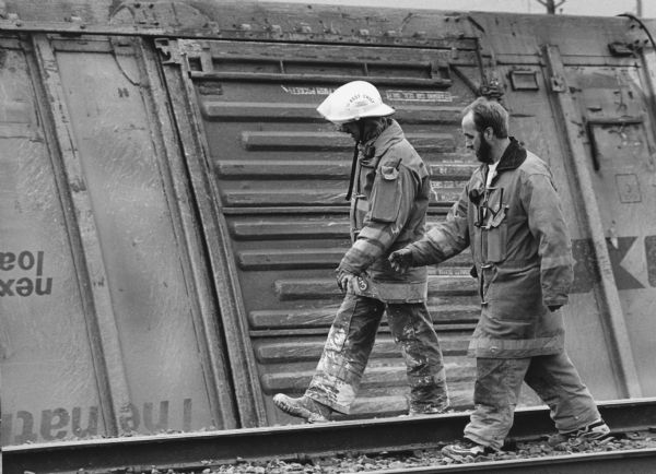 "First assistant fire chief Chuck Trauba and fireman, Dale Krueger, surveyed the damage along the tracks with an overturned railcar as a backdrop."