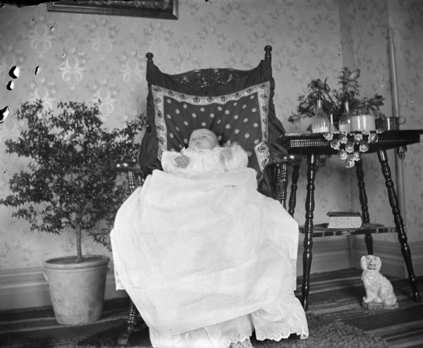 A baby portrait of Edgar Krueger at age 2 months lying in a chair wearing a long gown. Bottles of milk are sitting on a side table.