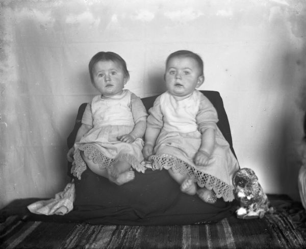 Indoor portrait of Edgar and Jennie Krueger, at age 11 months and 17 days, posing in front of a light cloth backdrop. They are wearing matching dresses with a crochet trim and are sitting on a seat or cushion draped with a dark cloth. A rug is on the floor. There is a doll on the floor on the left, and a toy animal on wheels is on the right.