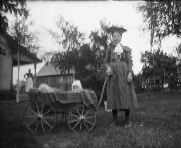Hattie Braemer, the children's caretaker, is standing next to the twins who are sitting in a wagon. A woman is standing on the porch behind them.