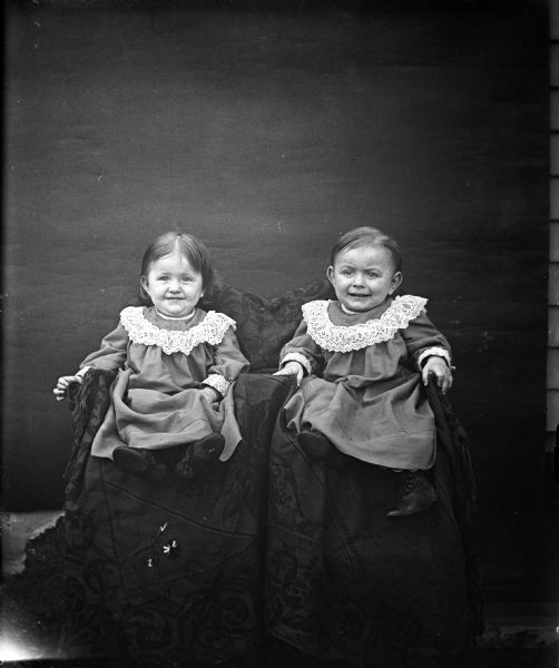 Portrait of the twins, Jennie and Edgar Krueger. They are wearing matching outfits and sitting in chairs draped in fabric.