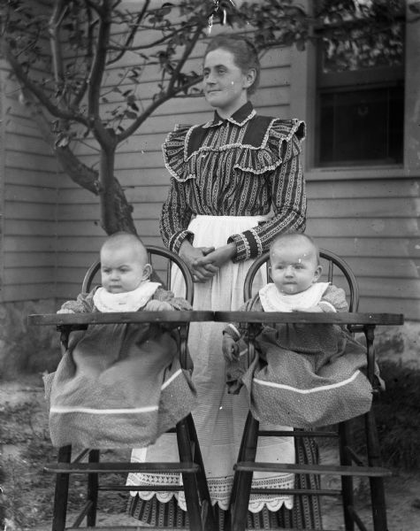 Flora Krueger is standing behind the twins, aged 6 months, 7 days, who are sitting in highchairs. Edgar, left, weighs 23 lbs., Jennie, right, weighs 19 lbs.