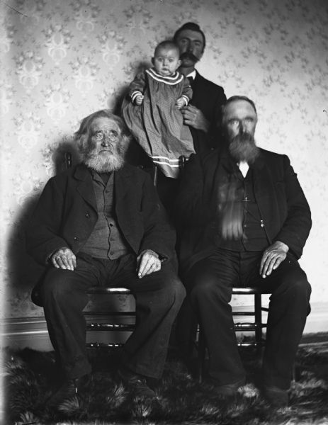 Portrait of four generations of Krueger men. William and August are sitting from left to right. Behind them, Alex is standing holding Edgar. There is a fur rug on the floor.